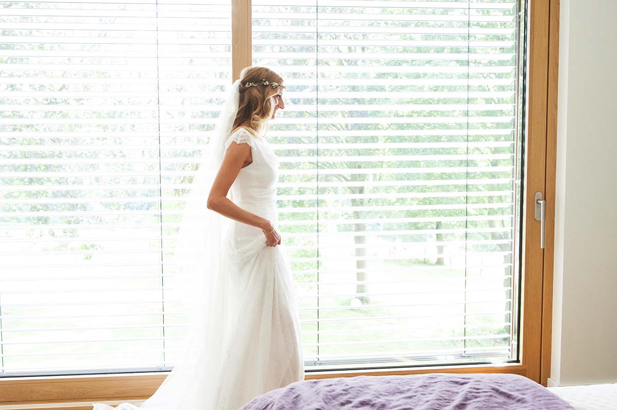 Honeylight Hochzeitsfotografen: Braut im Brautkleid in Hochzeitsvorbereitung bei der Anprobe vor dem Fenster