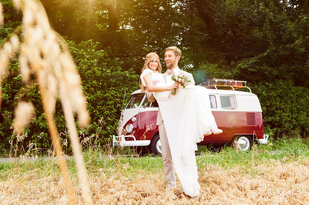 Honeylight Hochzeitsfotografen: Bräutigam trägt die Braut auf einem Feld. Im Hintergrund ein roter VW Bulli